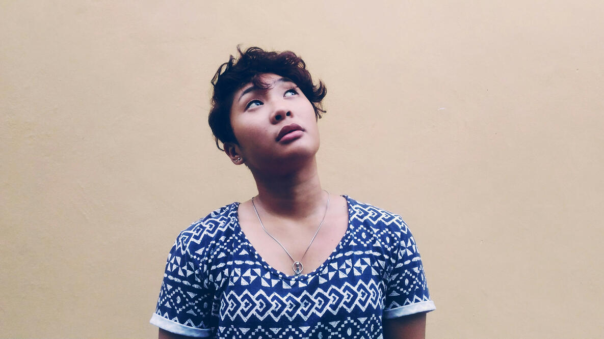 A female-presenting person with short, wavy hair and wearing a blue patterned shirt looks upward with a contemplative expression. They are standing against a plain beige wall. The image, by Mel Lituanas for Unsplash, conveys a sense of thoughtfulness.
